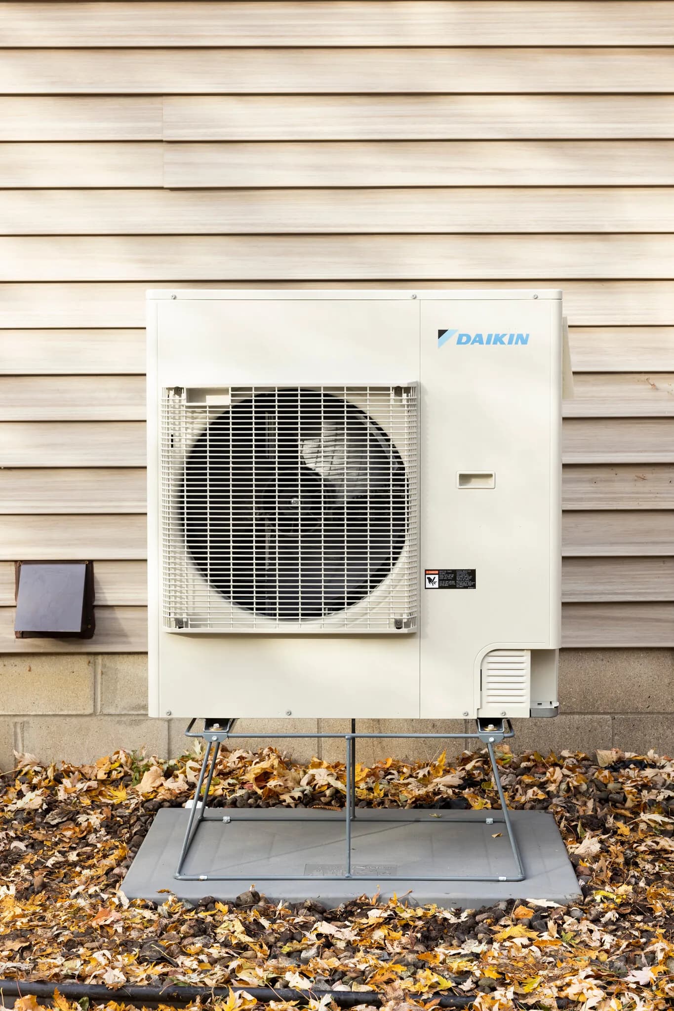 Daikin heat pump outdoor unit mounted on a raised stand against siding, surrounded by fall leaves
