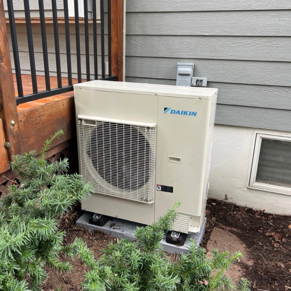 Daikin heat pump outdoor unit installed beside a wooden deck with evergreen shrubs