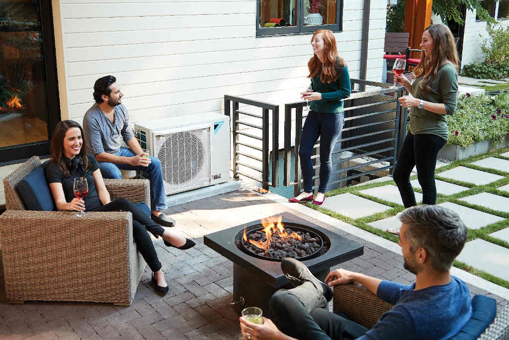 Friends relaxing around a patio fire pit beside a white Daikin FIT heat pump against the home siding